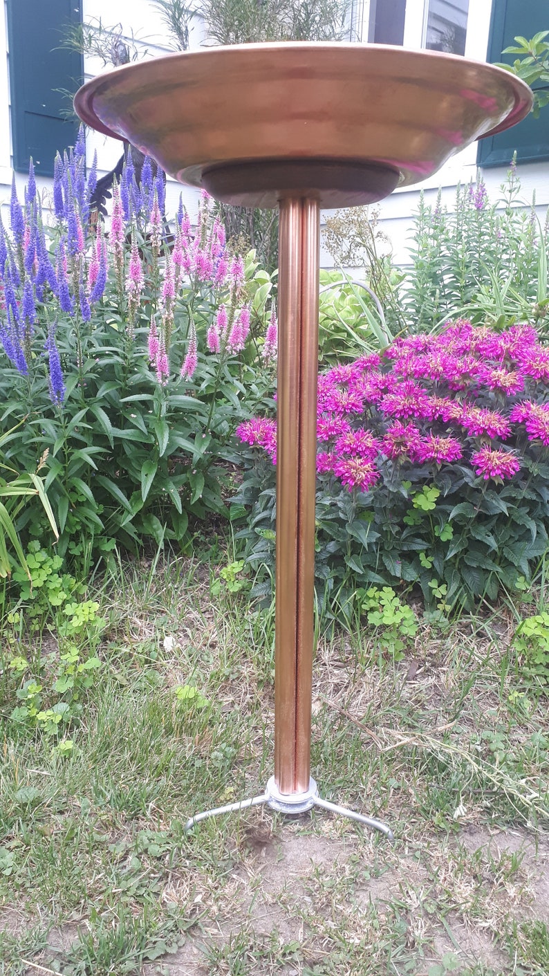 May include: A copper bird bath with a wide, shallow bowl and a tall, slender stand. The stand has three legs that extend out from the base. The bird bath is surrounded by green grass and pink and purple flowers.