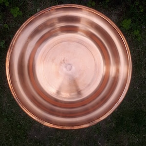 May include: A round copper bird bath with a slightly raised rim. The bird bath is sitting on a bed of green grass.