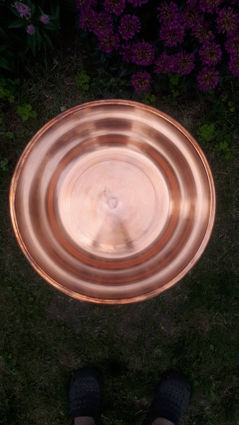 May include: A round copper bird bath with a slightly raised rim. The bird bath is sitting on a bed of green grass.