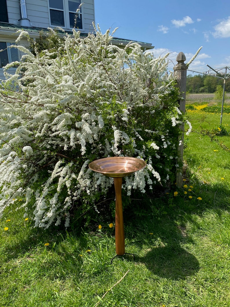 May include: A copper bird bath on a wooden stand with a three-legged base. The bird bath is in a grassy area with a bush of white flowers in the background.