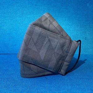May include: A gray face mask with a geometric pattern. The mask has a black elastic band.