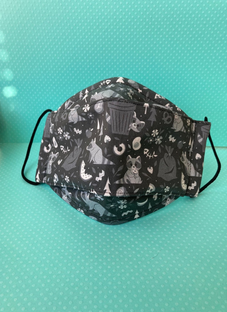 K&ouml;nnte beinhalten: Schwarze Gesichtsmaske mit einem wei&szlig;en Muster aus Waschb&auml;ren, B&auml;umen und M&uuml;lleimern.