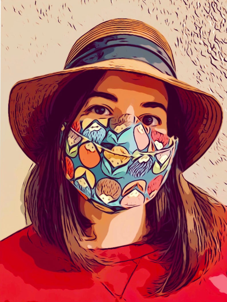 K&ouml;nnte beinhalten: Eine Frau tr&auml;gt einen Strohhut mit einem blauen Band und eine blaue und gr&uuml;ne gemusterte Gesichtsmaske mit bunten Fr&uuml;chten und Blumenmustern.