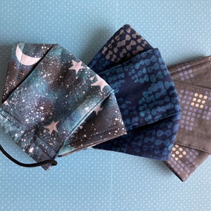 K&ouml;nnte beinhalten: Drei Stoffmasken mit verschiedenen Mustern. Eine Maske hat einen blauen und schwarzen Galaxie-Druck mit wei&szlig;en Sternen und einem Halbmond. Eine weitere Maske hat ein dunkelblaues und hellblaues Muster mit kleinen wei&szlig;en Punkten. Die dritte Maske hat ein dunkelgraues und hellgraues Muster mit kleinen wei&szlig;en Punkten.