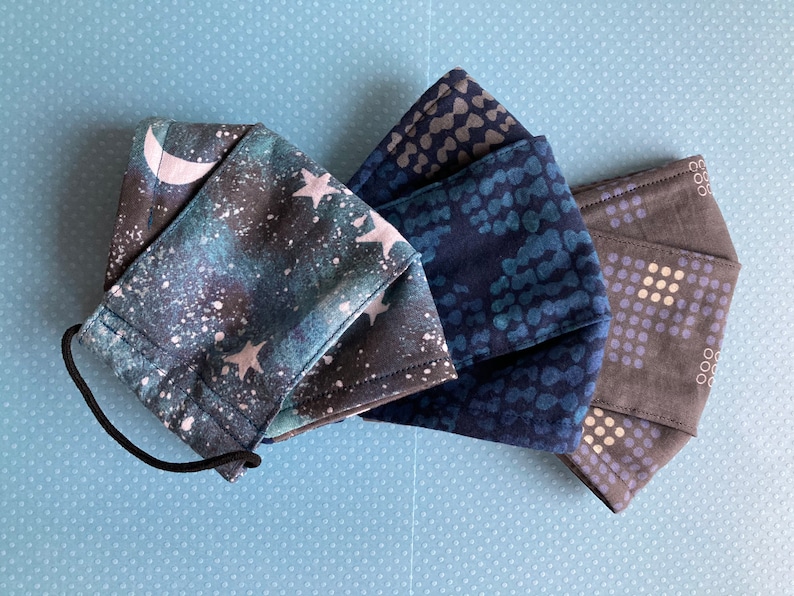 K&ouml;nnte beinhalten: Drei Stoffmasken mit verschiedenen Mustern. Eine Maske hat einen blauen und schwarzen Galaxie-Druck mit wei&szlig;en Sternen und einem Halbmond. Eine weitere Maske hat ein dunkelblaues und hellblaues Muster mit kleinen wei&szlig;en Punkten. Die dritte Maske hat ein dunkelgraues und hellgraues Muster mit kleinen wei&szlig;en Punkten.