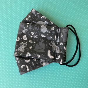 K&ouml;nnte beinhalten: Eine schwarze Gesichtsmaske mit einem wei&szlig;en Muster aus Waschb&auml;ren, M&uuml;lleimern, Wassermelonen und anderen Waldmotiven. Die Maske hat schwarze elastische Ohrschlaufen.