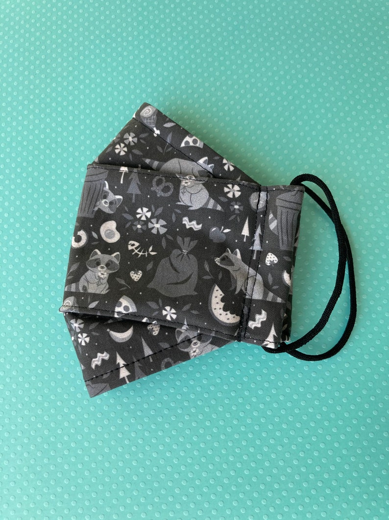 K&ouml;nnte beinhalten: Eine schwarze Gesichtsmaske mit einem wei&szlig;en Muster aus Waschb&auml;ren, M&uuml;lleimern, Wassermelonen und anderen Waldmotiven. Die Maske hat schwarze elastische Ohrschlaufen.