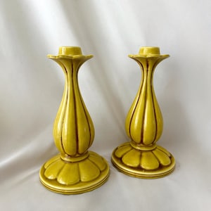 May include: A pair of vintage ceramic candle holders in a mustard yellow hue. Each candle holder features a scalloped base, a bulbous body, and a flared top. The candle holders are approximately 20 cm tall and have a glossy finish.
