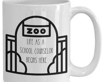 May include: White ceramic mug with a black graphic of a zoo entrance and the text "ZOO" above "LIFE AS A SCHOOL COUNSELOR BEGINS HERE". The mug has a curved handle.