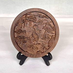 May include: A round, wooden decorative plate with a detailed relief carving of a crane, mountains, and clouds. The plate is brown and has a decorative border. It is displayed on a black stand.