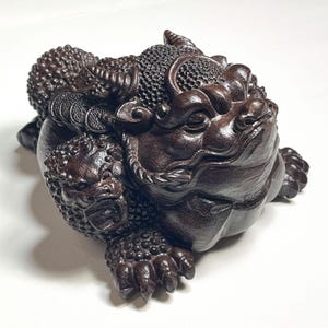 Puede incluir: Escultura de madera marrón oscuro de un sapo de tres patas, un símbolo tradicional de riqueza. El sapo tiene tallas detalladas, incluyendo monedas y una pequeña figura de guardián. Sobre un fondo blanco.