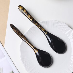 May include: Two black spoons with a glossy finish and gold-toned, circular, organic patterns along the handles. The spoons feature a deep, rounded bowl shape and a long, slender handle, resting on a white, scalloped-edge plate.