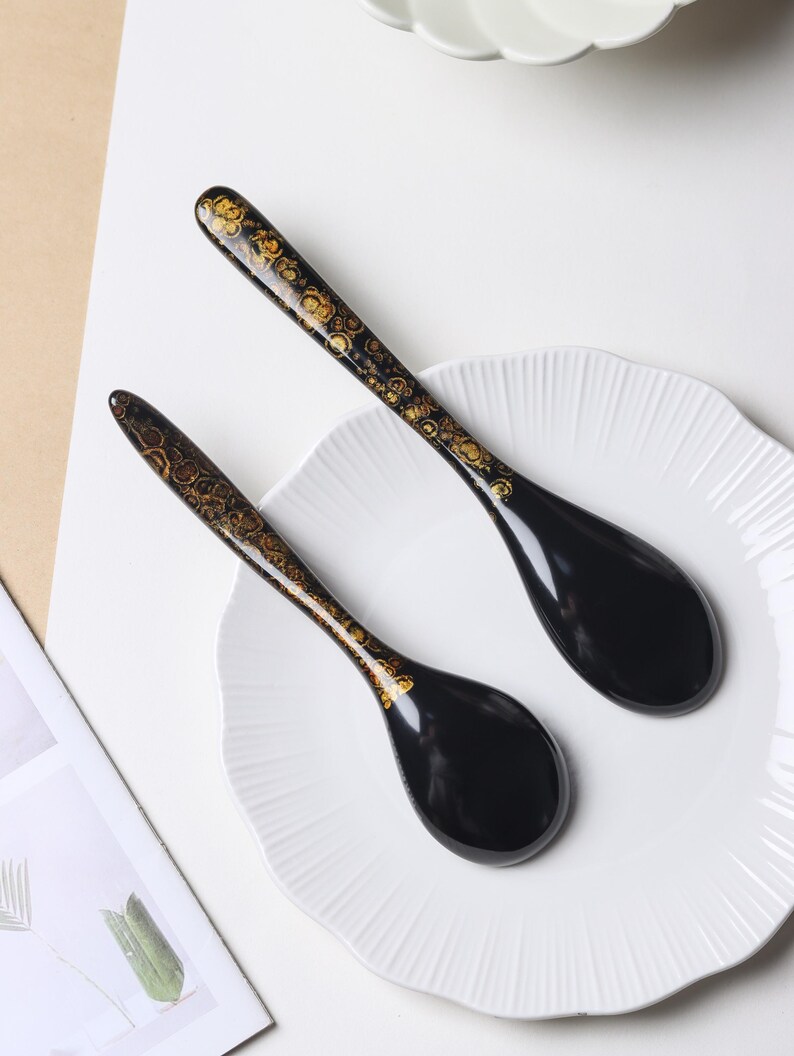 May include: Two black spoons with a glossy finish and gold-toned, circular, organic patterns along the handles. The spoons feature a deep, rounded bowl shape and a long, slender handle, resting on a white, scalloped-edge plate.