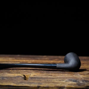 May include: A black tobacco pipe with a curved bowl and a long, straight stem rests on a wooden surface. The pipe is made of dark material, and the background is a solid black.