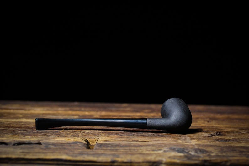 May include: A black tobacco pipe with a curved bowl and a long, straight stem rests on a wooden surface. The pipe is made of dark material, and the background is a solid black.
