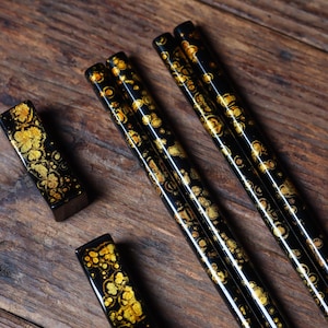 May include: A set of four black chopsticks with gold flecks. The chopsticks are arranged in a row on a wooden surface. There are two matching chopstick rests with the same gold fleck design.