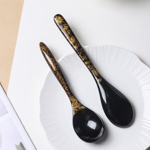 May include: Two black soup spoons with long, slender handles featuring a decorative gold and brown swirling pattern. The bowls of the spoons are smooth, solid black, and glossy. The spoons are arranged diagonally on a white, scalloped-edge plate.