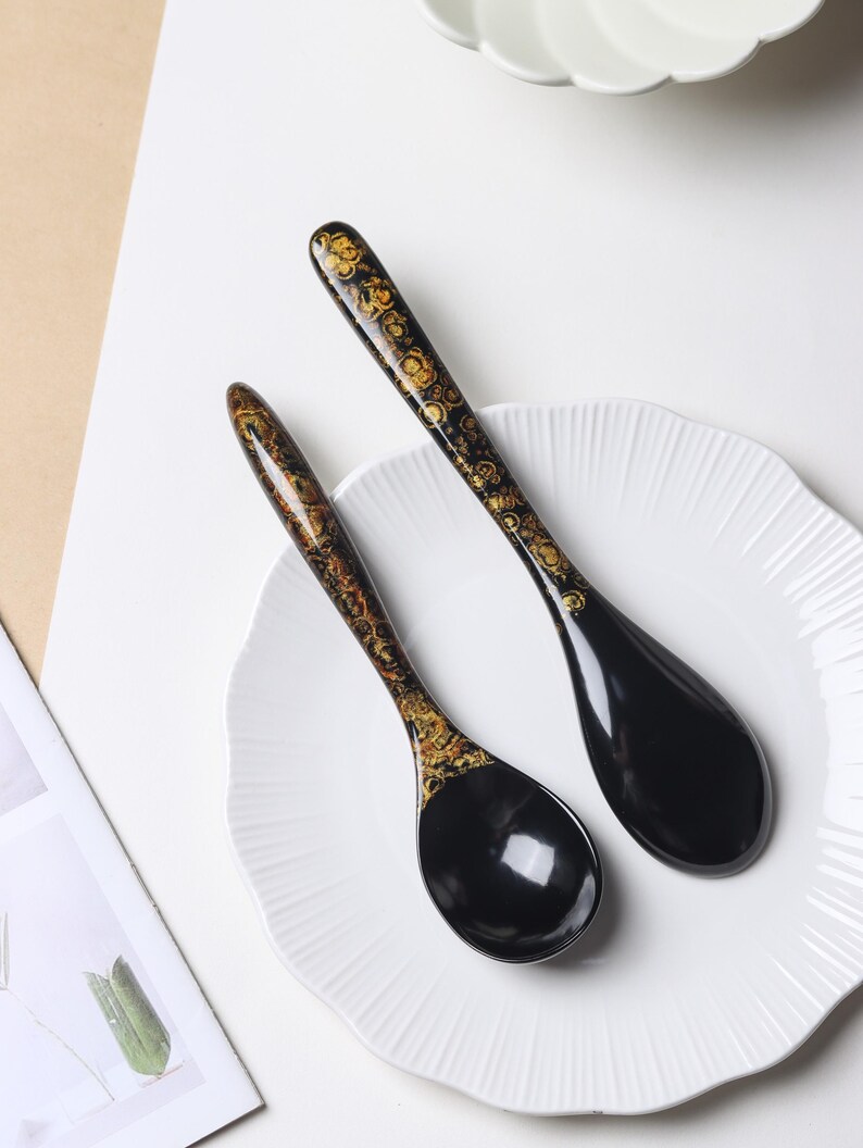 May include: Two black soup spoons with long, slender handles featuring a decorative gold and brown swirling pattern. The bowls of the spoons are smooth, solid black, and glossy. The spoons are arranged diagonally on a white, scalloped-edge plate.