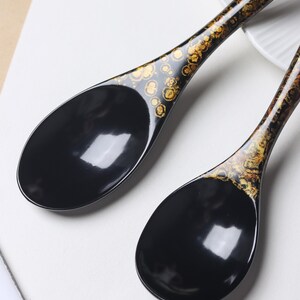 May include: Two black soup spoons with a glossy finish. The handles feature a decorative pattern of gold-colored, circular, and organic-shaped flecks that transition into the solid black, deep, oval-shaped bowls of the spoons.