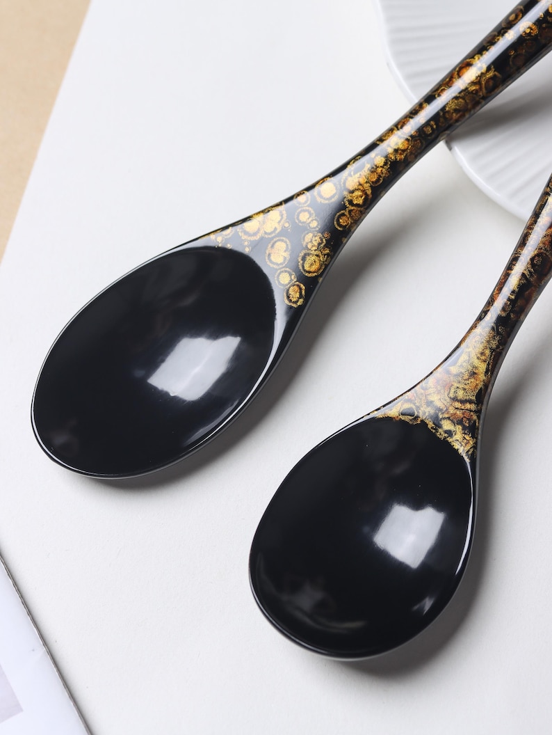 May include: Two black soup spoons with a glossy finish. The handles feature a decorative pattern of gold-colored, circular, and organic-shaped flecks that transition into the solid black, deep, oval-shaped bowls of the spoons.