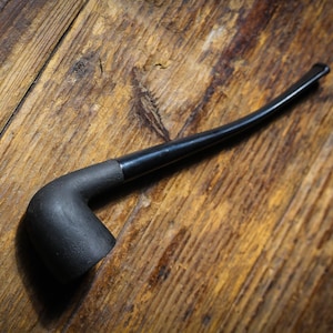 May include: A black smoking pipe with a curved stem lies on a wooden surface. The pipe features a dark, matte bowl and a glossy black stem. The wooden background provides a rustic aesthetic.