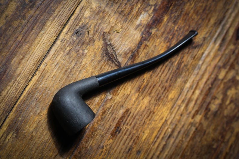 May include: A black smoking pipe with a curved stem lies on a wooden surface. The pipe features a dark, matte bowl and a glossy black stem. The wooden background provides a rustic aesthetic.