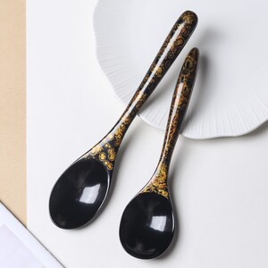 May include: Two black soup spoons with long, slender handles featuring a decorative gold and brown mottled pattern. The bowls of the spoons are smooth, solid black, and have a glossy finish. The spoons are arranged diagonally on a white surface.