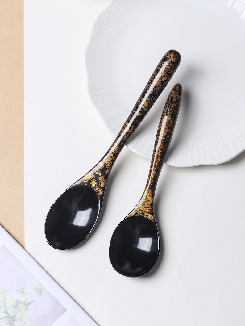 May include: Two black soup spoons with long, slender handles featuring a decorative gold and brown mottled pattern. The bowls of the spoons are smooth, solid black, and have a glossy finish. The spoons are arranged diagonally on a white surface.