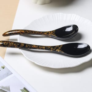 May include: Two black spoons with a glossy finish and a decorative gold-colored pattern featuring circular motifs along the handles. The spoons have a curved shape and are positioned on a white plate with a scalloped edge and a ribbed texture.
