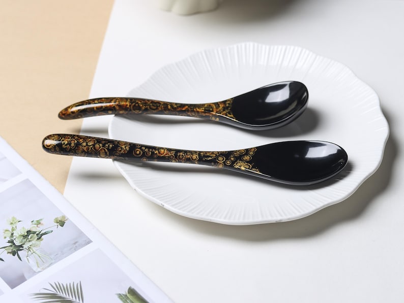 May include: Two black spoons with a glossy finish and a decorative gold-colored pattern featuring circular motifs along the handles. The spoons have a curved shape and are positioned on a white plate with a scalloped edge and a ribbed texture.