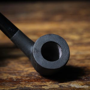 May include: A black smoking pipe, viewed close-up. The pipe features a curved stem and a circular bowl with a central opening. It is set against a wooden background. The pipe is made of a dark material.