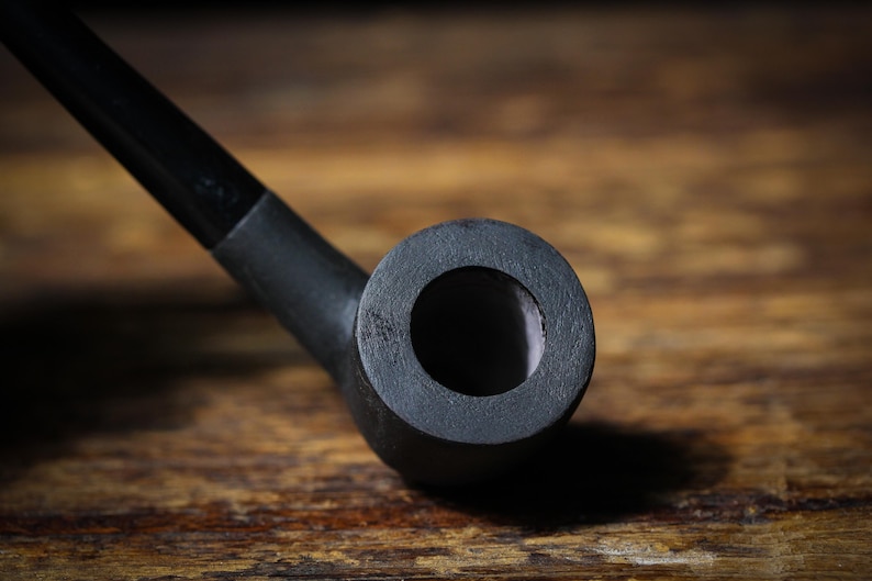 May include: A black smoking pipe, viewed close-up. The pipe features a curved stem and a circular bowl with a central opening. It is set against a wooden background. The pipe is made of a dark material.