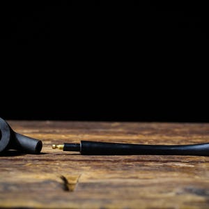 May include: A black tobacco pipe, in two parts, lies on a wooden surface against a black background. The pipe has a bowl, stem, and a brass fitting. The wood grain is visible.