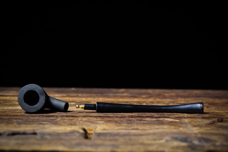 May include: A black tobacco pipe, in two parts, lies on a wooden surface against a black background. The pipe has a bowl, stem, and a brass fitting. The wood grain is visible.