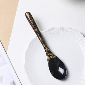 May include: A black spoon with a glossy finish featuring a decorative gold pattern of circular shapes along the handle and the upper portion of the bowl. The spoon has a long, slender handle and a wide, rounded bowl, resting on a white plate.
