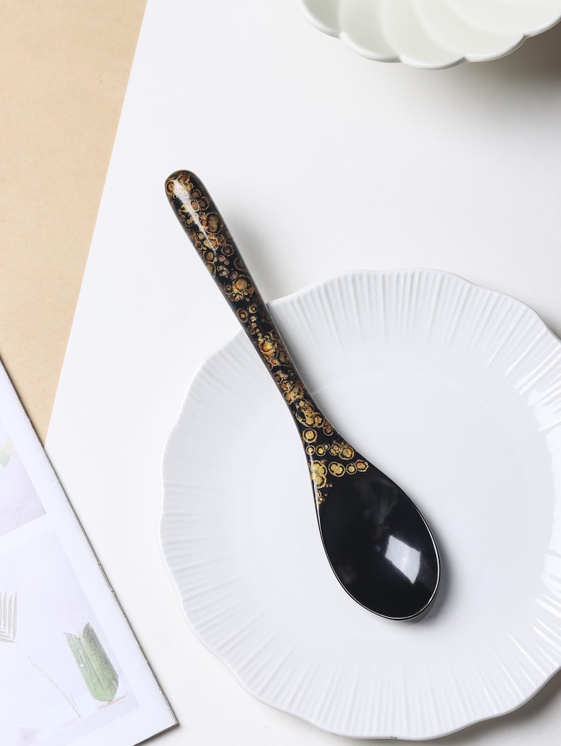 May include: A black spoon with a glossy finish featuring a decorative gold pattern of circular shapes along the handle and the upper portion of the bowl. The spoon has a long, slender handle and a wide, rounded bowl, resting on a white plate.