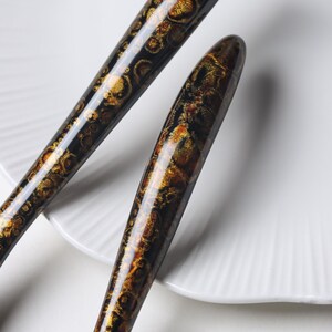 May include: Two elongated, tapered handles with a glossy finish, featuring a dark black base color overlaid with intricate, shimmering gold and bronze circular patterns that resemble organic, celestial, or mineral-like textures.