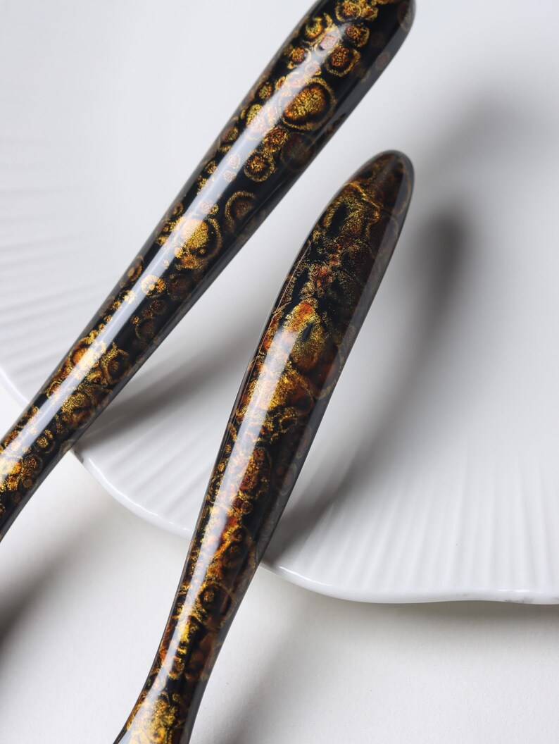 May include: Two elongated, tapered handles with a glossy finish, featuring a dark black base color overlaid with intricate, shimmering gold and bronze circular patterns that resemble organic, celestial, or mineral-like textures.