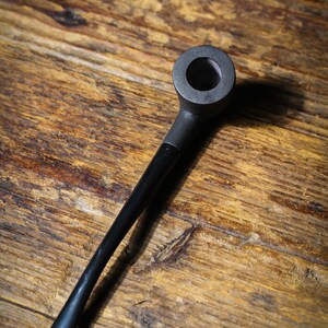 May include: A black smoking pipe with a rounded bowl and a long, slender stem. The pipe is made of a dark material, possibly wood or a composite, and rests on a wooden surface with visible grain.