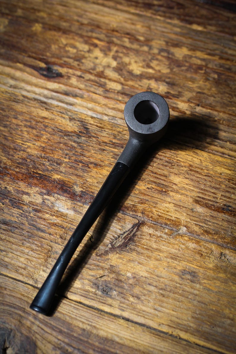 May include: A black smoking pipe with a rounded bowl and a long, slender stem. The pipe is made of a dark material, possibly wood or a composite, and rests on a wooden surface with visible grain.