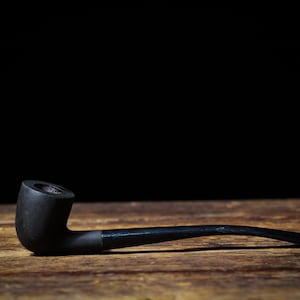 May include: A black smoking pipe with a curved stem, lying on a wooden surface. The pipe's bowl is cylindrical, and the stem tapers to a narrow mouthpiece. The background is black.