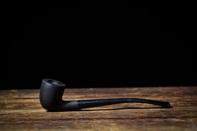 May include: A black smoking pipe with a curved stem, lying on a wooden surface. The pipe's bowl is cylindrical, and the stem tapers to a narrow mouthpiece. The background is black.