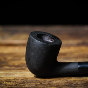 May include: A black smoking pipe with a curved stem and bowl, set on a wooden surface. The pipe's bowl is cylindrical, and the stem is smooth. The background is dark, highlighting the pipe's shape.