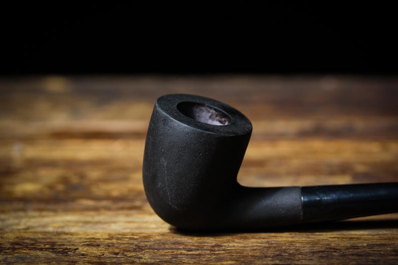 May include: A black smoking pipe with a curved stem and bowl, set on a wooden surface. The pipe's bowl is cylindrical, and the stem is smooth. The background is dark, highlighting the pipe's shape.