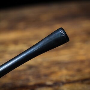 May include: Close-up of a black pipe stem, angled diagonally. The stem has a glossy finish and a tapered design, with a slightly flared end. The background is a blurred wooden surface.