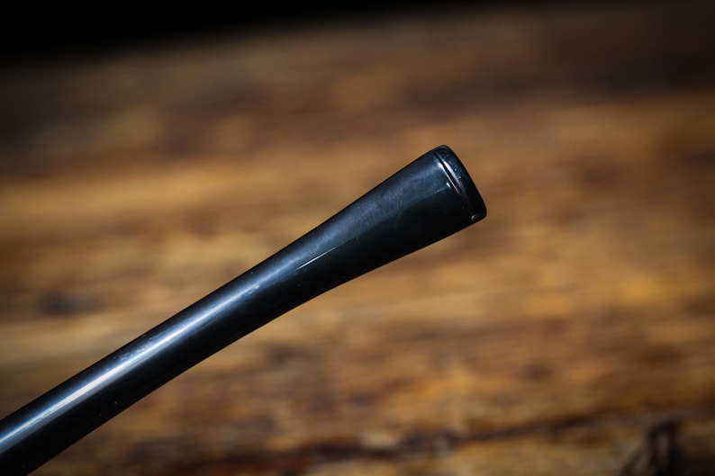 May include: Close-up of a black pipe stem, angled diagonally. The stem has a glossy finish and a tapered design, with a slightly flared end. The background is a blurred wooden surface.