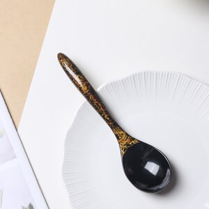 May include: A glossy spoon featuring a solid black bowl and a handle decorated with a mottled black and gold pattern. The spoon is positioned diagonally across a white plate with a scalloped edge, showcasing a smooth, reflective finish.