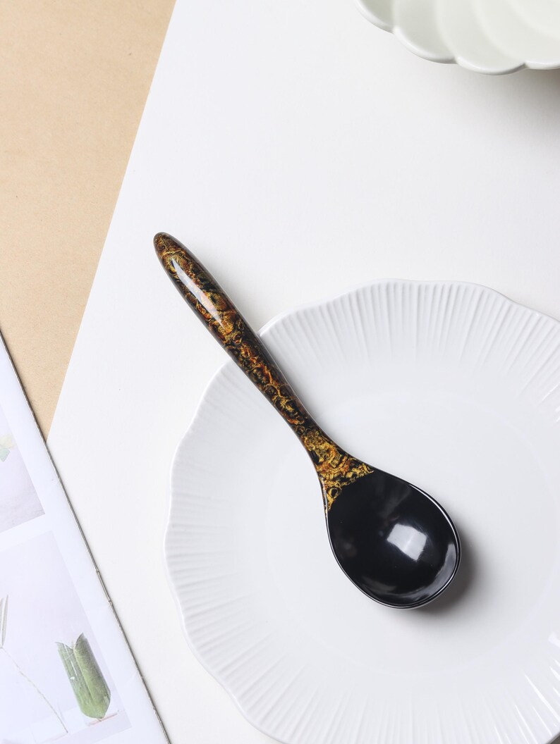 May include: A glossy spoon featuring a solid black bowl and a handle decorated with a mottled black and gold pattern. The spoon is positioned diagonally across a white plate with a scalloped edge, showcasing a smooth, reflective finish.