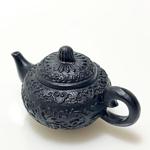 May include: A small, black teapot with intricate floral and scrollwork carvings. The teapot features a curved spout, a rounded body, and a loop handle. The lid is adorned with a decorative knob.