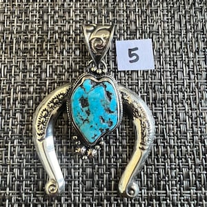 May include: A silver pendant featuring a central irregular turquoise stone with black matrix, set within a horseshoe-shaped frame with engraved floral patterns and small silver beads. The bail has an etched design and a white tag with the number 5.